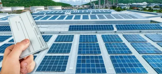 A person is holding a check in front of a roof with solar panels.