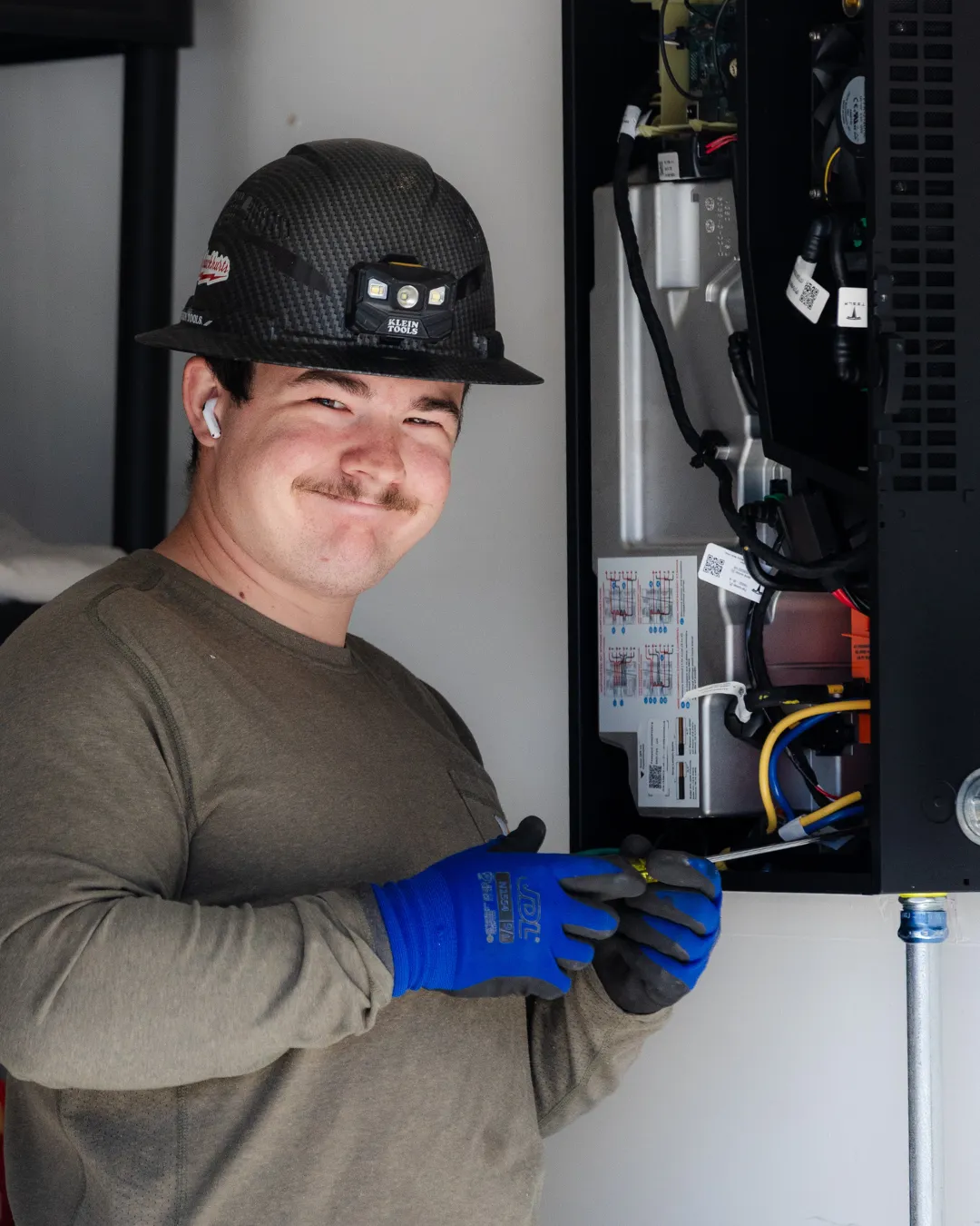 Technician in hard hat and blue gloves working on exposed electrical wiring in a cabinet.