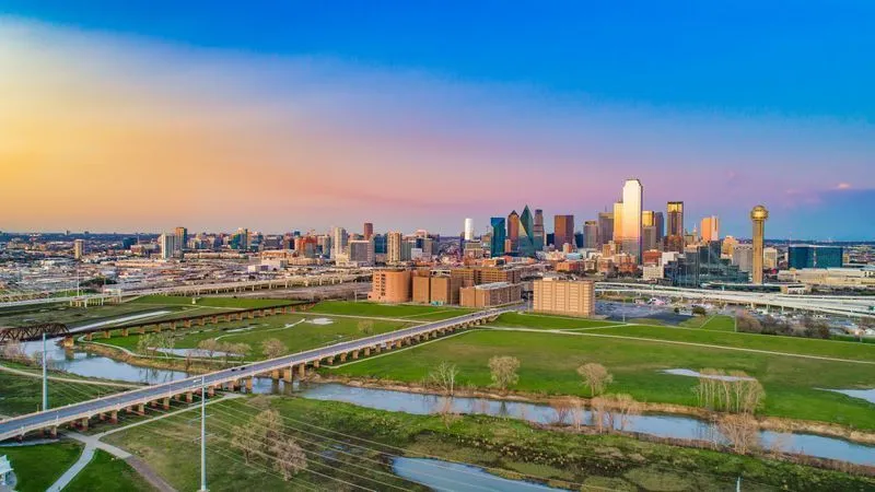 City skyline at sunset over green parkland and winding waterways under a colorful sky