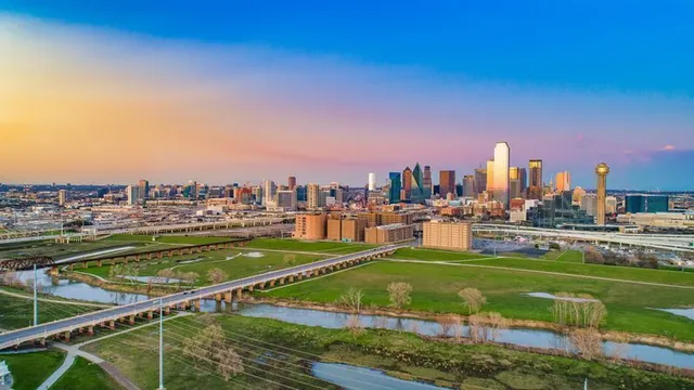 City skyline at sunset over green parkland and winding waterways under a colorful sky