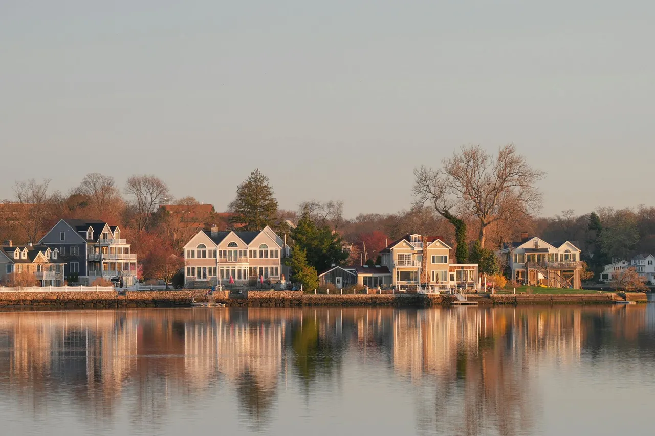 Row of waterfront houses reflected in calm water at sunset, with bare trees along the shore