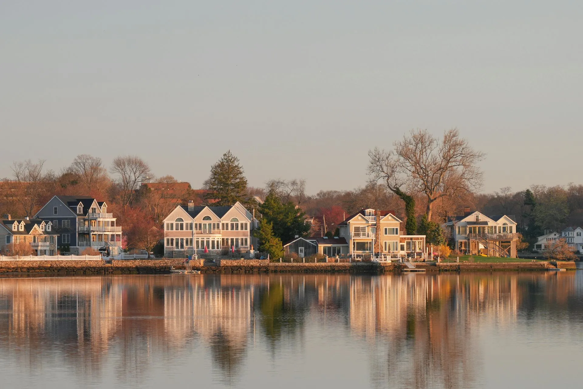 Row of waterfront houses reflected in calm water at sunset, with bare trees along the shore