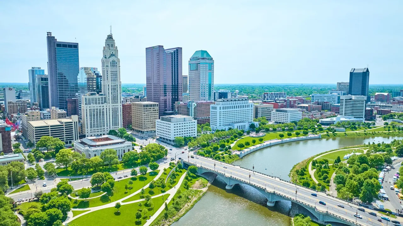 Aerial cityscape with modern high-rises, a winding river, bridge, and green parks under a clear sky.
