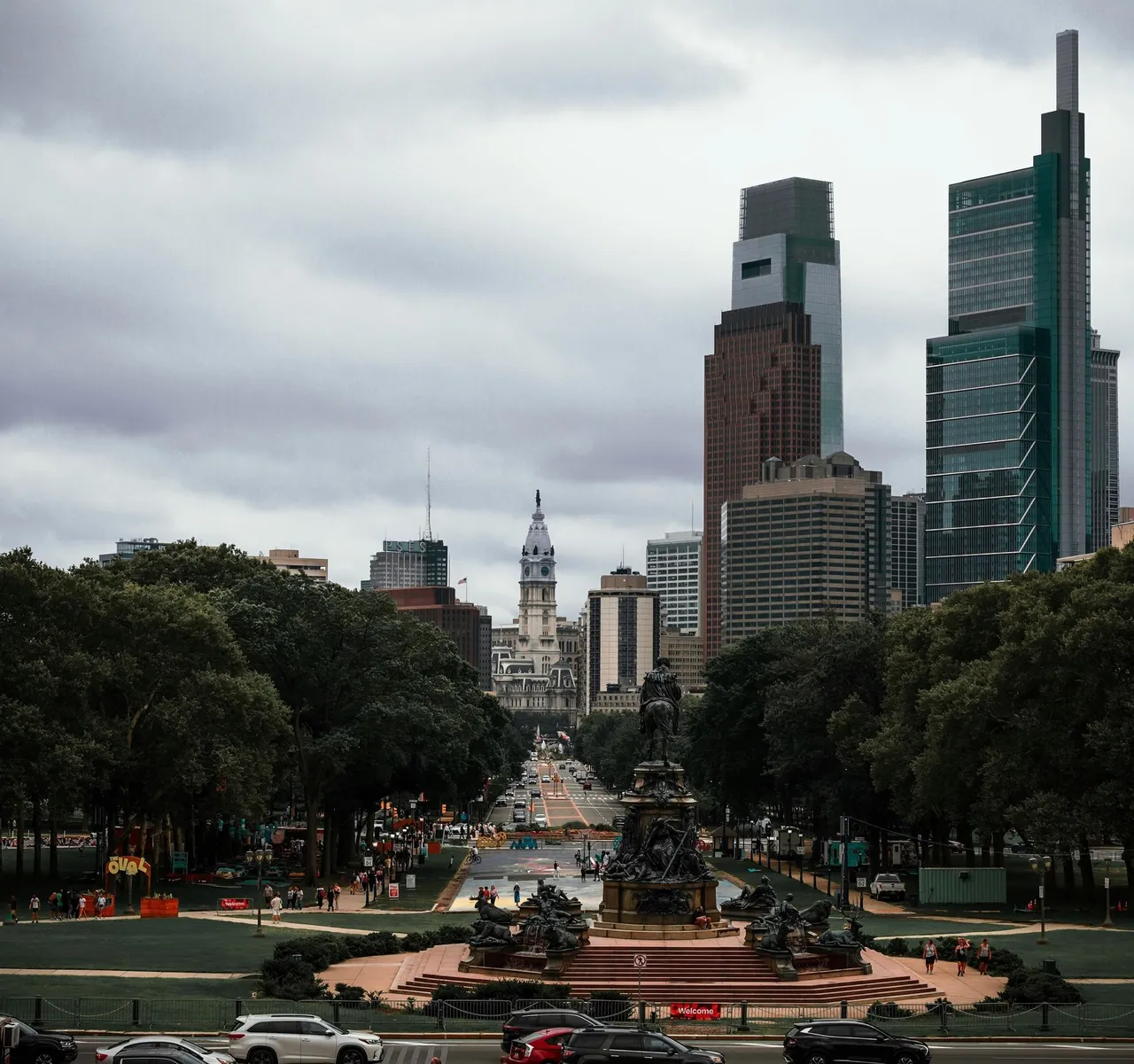 City skyline framed by trees, with a central monument and cloudy sky over a broad boulevard.