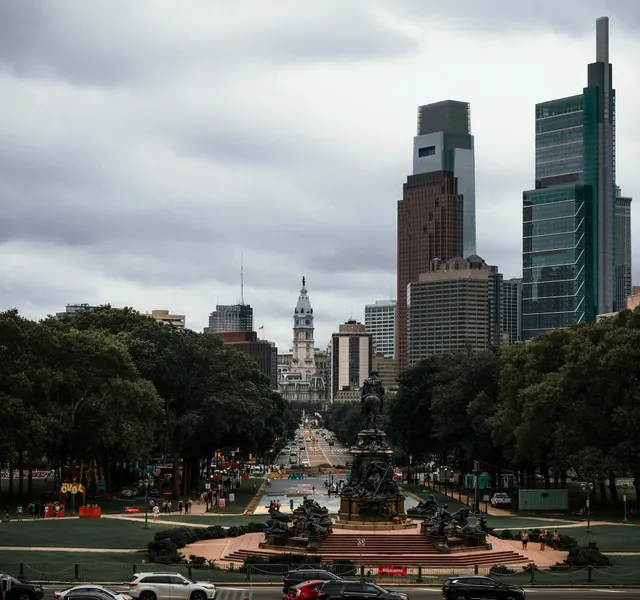 City skyline framed by trees, with a central monument and cloudy sky over a broad boulevard.