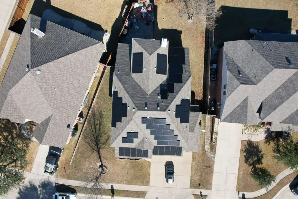 An aerial view of a house with solar panels on the roof Aerial view of three suburban houses with dark gray roofs and parked cars on driveways.