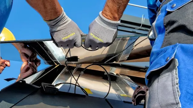 Gloved hands working on cracked black glass or windshield outdoors under a blue sky.