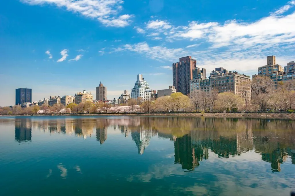 City skyline reflected in a calm lake under a blue sky with scattered clouds.